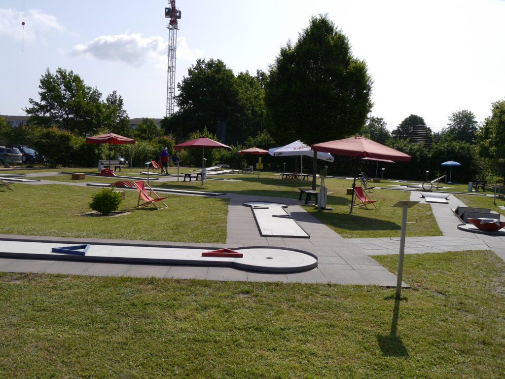 Minigolfanlage bei Sonnenschein mit Sonnenschirmen und Klappstühlen zwischen den Bahnen.
