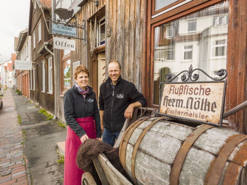 Zwei Personen stehen vor dem Laden des Fischereibetriebs Nölke und einem antiken Ladenschild.