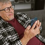 Ein älterer Mann mit Brille sitzt auf dem Sofa und bedient ein Smartphone
