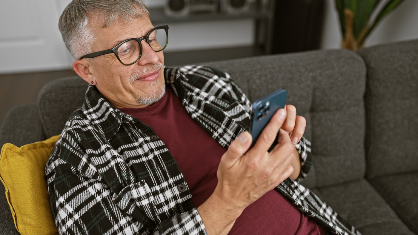 Ein älterer Mann mit Brille sitzt auf dem Sofa und bedient ein Smartphone
