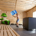 Das Frosch-Maskottchen der Stadtwerke Celle sitzt allein in einer Sauna und bekommt Luft mit einem Handtuch zugefächert.